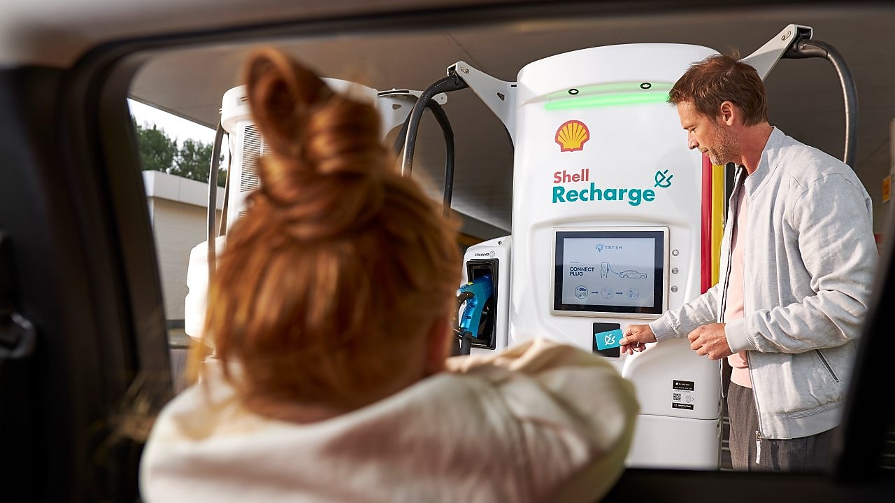 A man using a Shell Recharge unit while a girl watches from inside a car