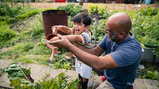 Man helping a smiling child water plants in a garden