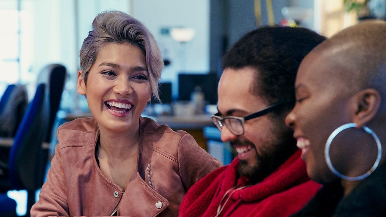 Three people in an office sitting together, looking at papers and laughing