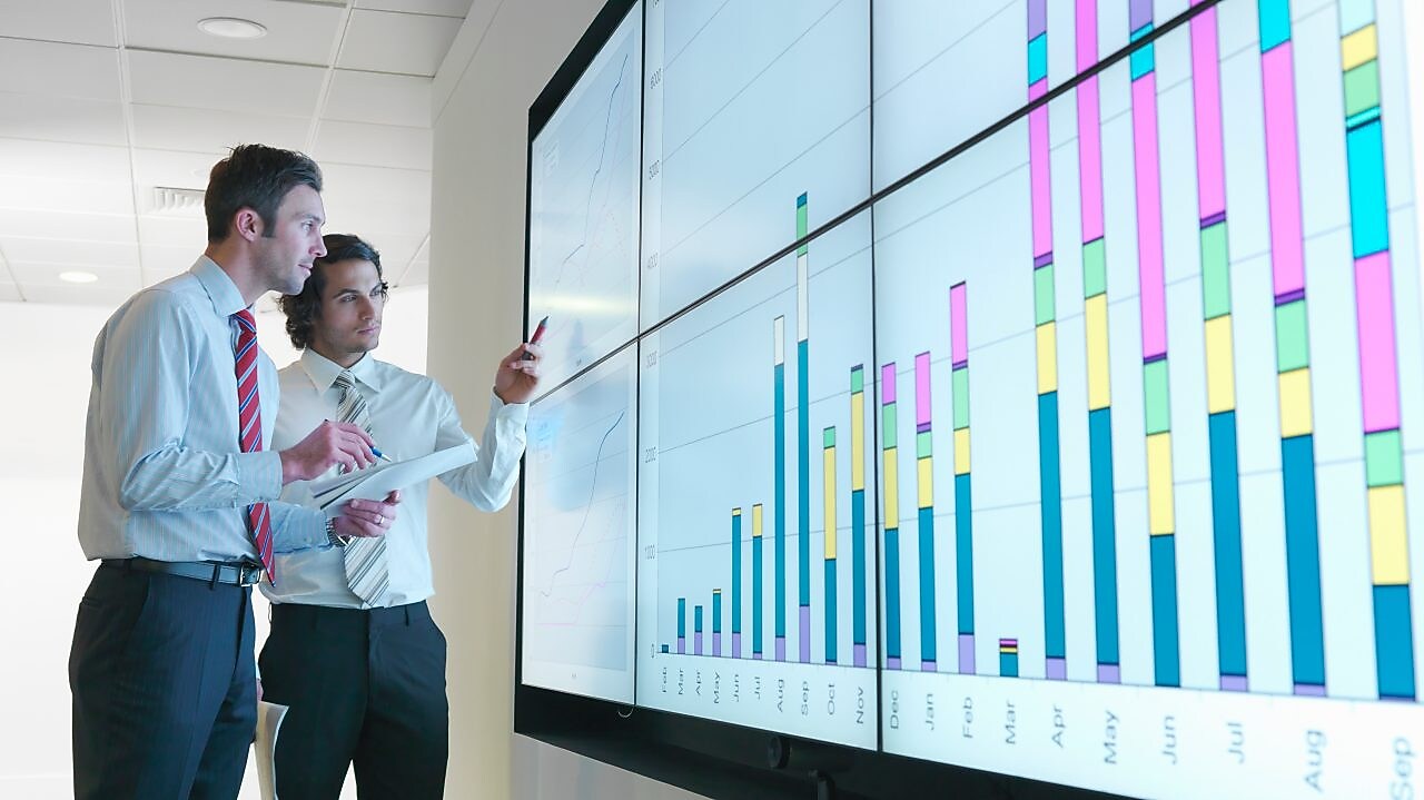 Business men analyse reports displayed on a large digital screen